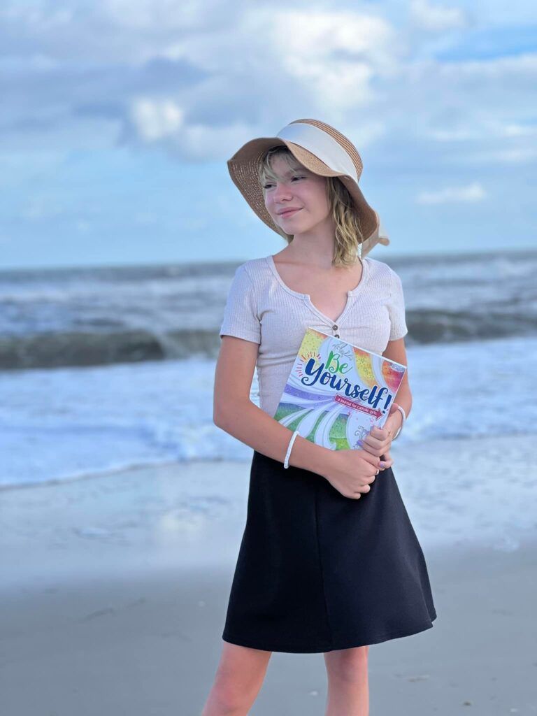 Young girl on beach holding a book titled Be Yourself